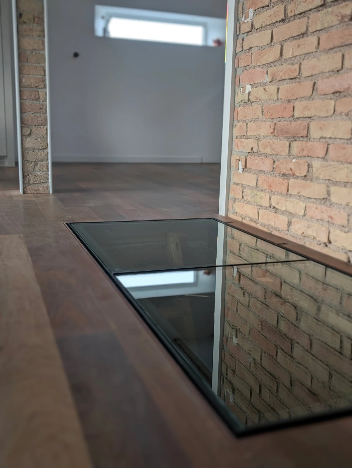 Trappe de cave vitrée à cadre noir intégrée dans un parquet, près d’un mur en briques — réalisation Art et Fer